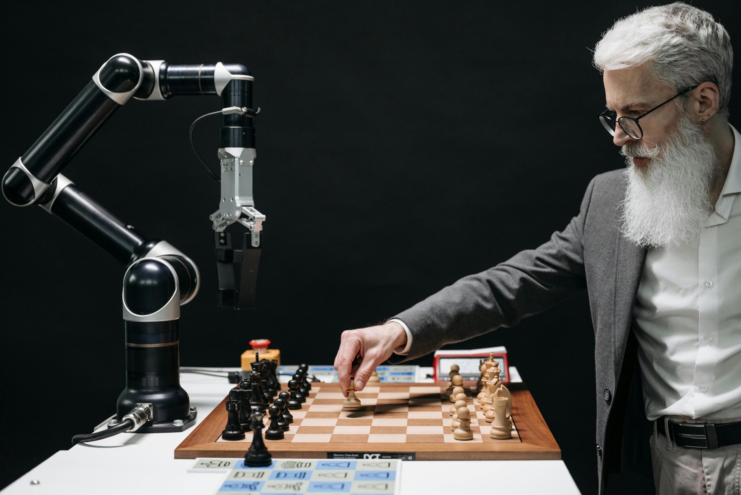 Man with white hair and beard in grey suit jacket playing chess with a robotic arm.