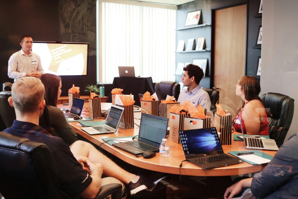 Group of people sitting around an oval conference table. Each has a laptop and a notebook. A facilitator is at the front of the room teaching them about something.