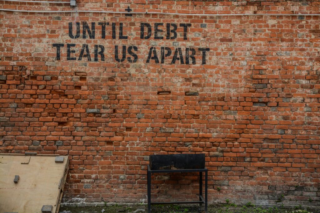 Red brick wall with black graffiti text reading “UNTIL DEBT TEAR US APART,” with a small black metal stand and a wooden board leaning nearby.