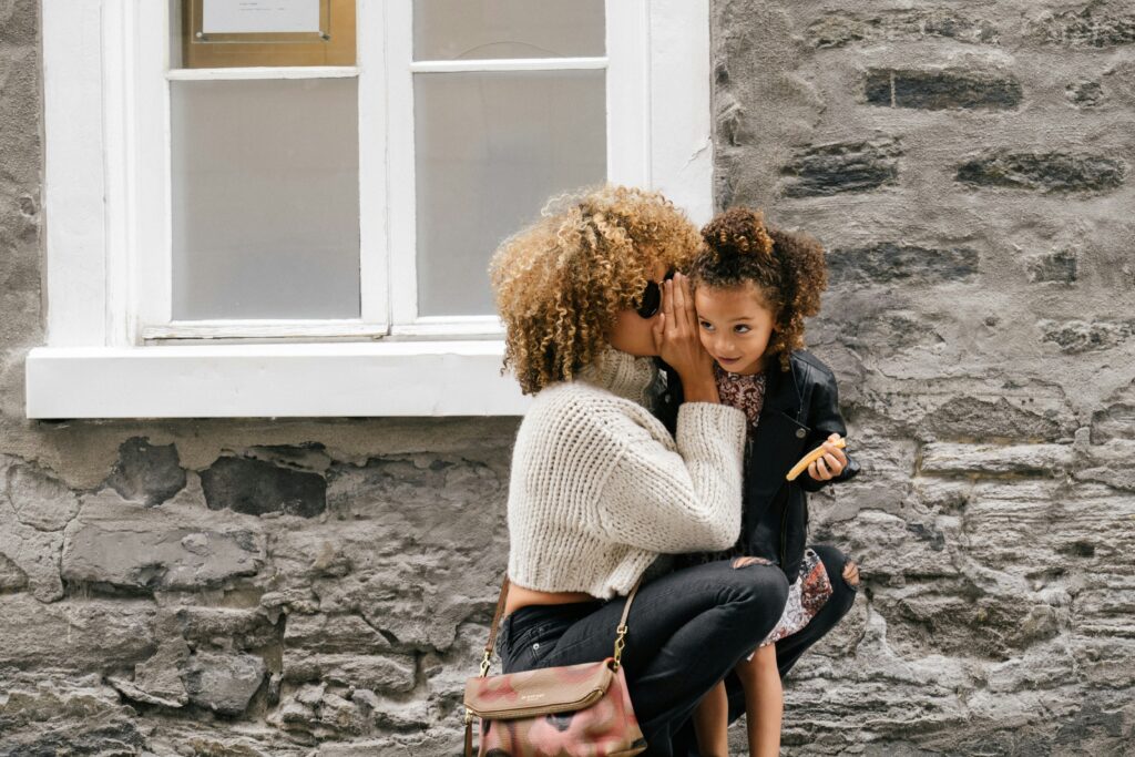 Woman whispering into the ear of a little girl who listening intently. They are in front of a stone house with a double clad white window.