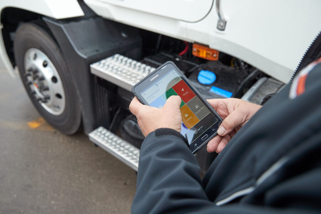 A truck driver stands beside a commercial vehicle using a Samsung tablet to access a fleet management app, showing route, vehicle, and driving data—illustrating Samsara’s mobile tools for monitoring and improving driver performance.