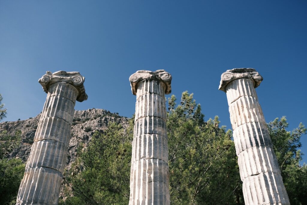 Three ancient Greek columns stand amid green trees with a rocky mountain backdrop under a bright blue sky.