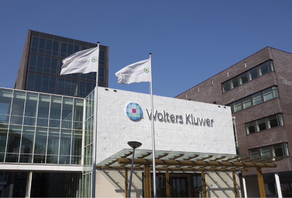 Exterior of the Wolters Kluwer headquarters showing the company logo on a white stone facade, two Wolters Kluwer flags flying, and modern glass and brick office buildings under a clear blue sky.

